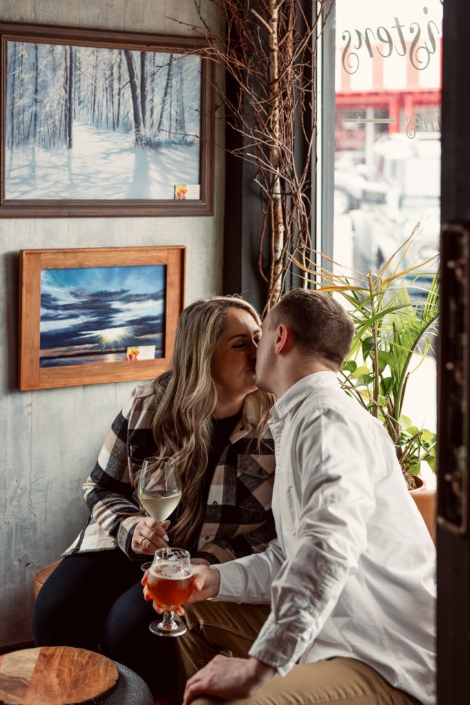 Bianca and Nick sharing a kiss in a cozy setting, holding drinks, with artwork on the walls, capturing their engagement moment in Hayward, WI.