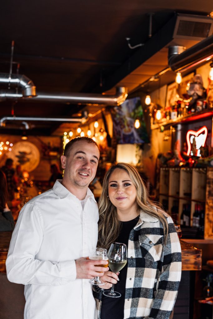 Bianca and Nick holding drinks in a cozy venue, celebrating their engagement in Hayward, WI.