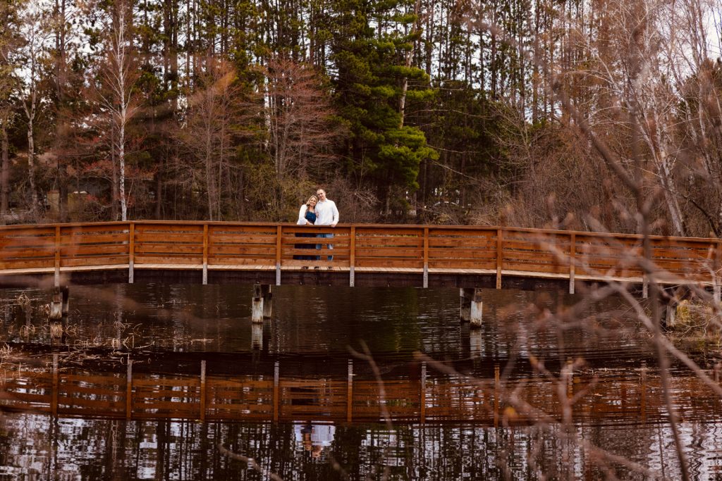 Bianca and Nick standing on a wooden bridge over a tranquil pond in Hayward, WI, surrounded by trees, capturing their engagement moment.