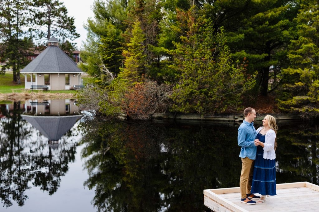 Couple embracing on a dock by a serene lake in Hayward, WI, with a gazebo and lush trees reflected in the water, capturing a romantic engagement moment.