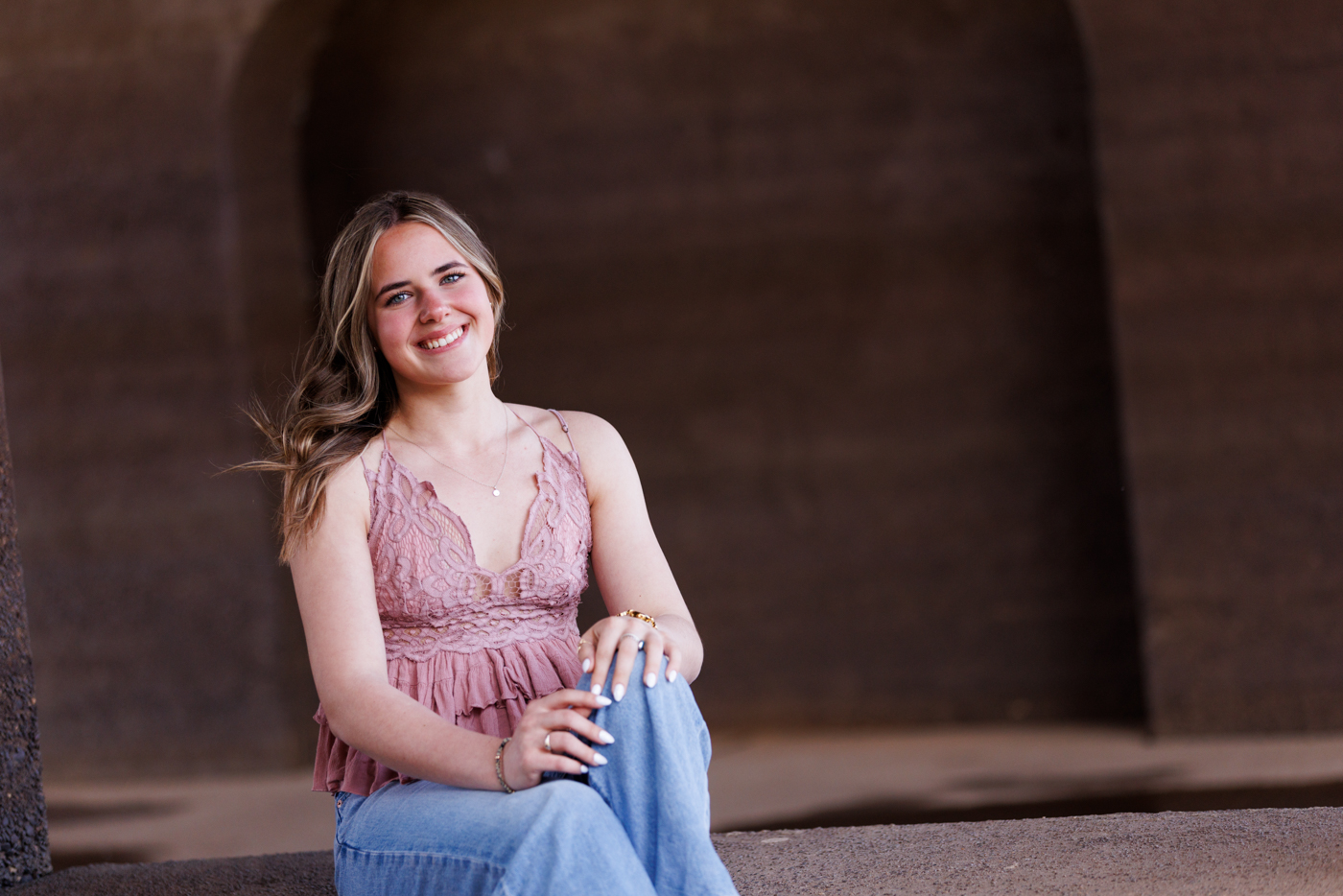 Senior portrait of a smiling young woman in a pink lace top and jeans, seated in a natural setting, highlighting personal style and warmth for portrait photography services.