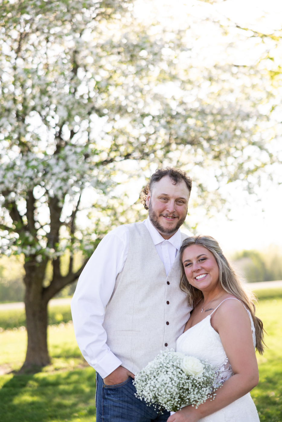 Couple smiling together at their wedding, surrounded by blooming trees, with the bride holding a bouquet of baby’s breath and a white rose, capturing a joyful moment in a natural setting.