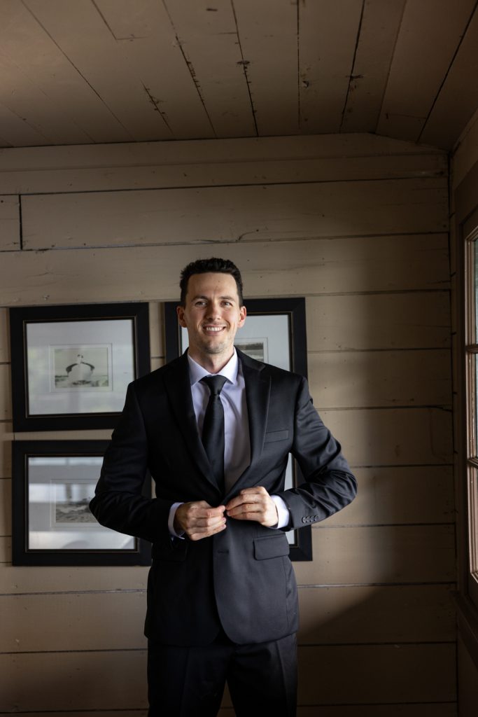 Groom in suit adjusting tie in front of framed photographs at Stout’s Island wedding venue