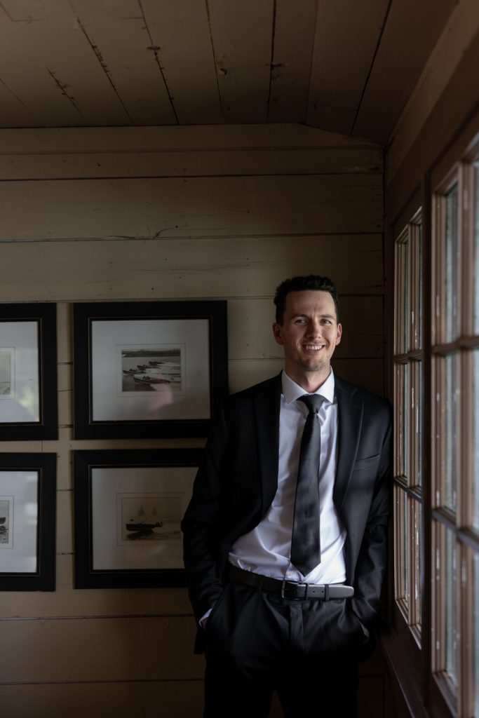 Groom in suit smiling near window and framed artwork at Stout’s Island Lodge wedding in