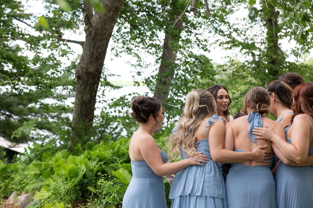 Bride and bridesmaids in blue dresses embracing near Red Cedar Lake at Stout’s Island wedding.