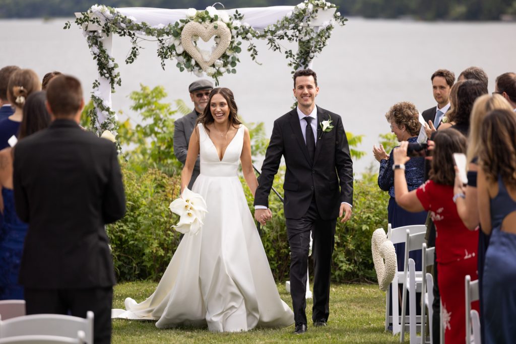 Brittany and Cole walking down aisle at Stout’s Island wedding ceremony, Red Cedar Lake backdrop