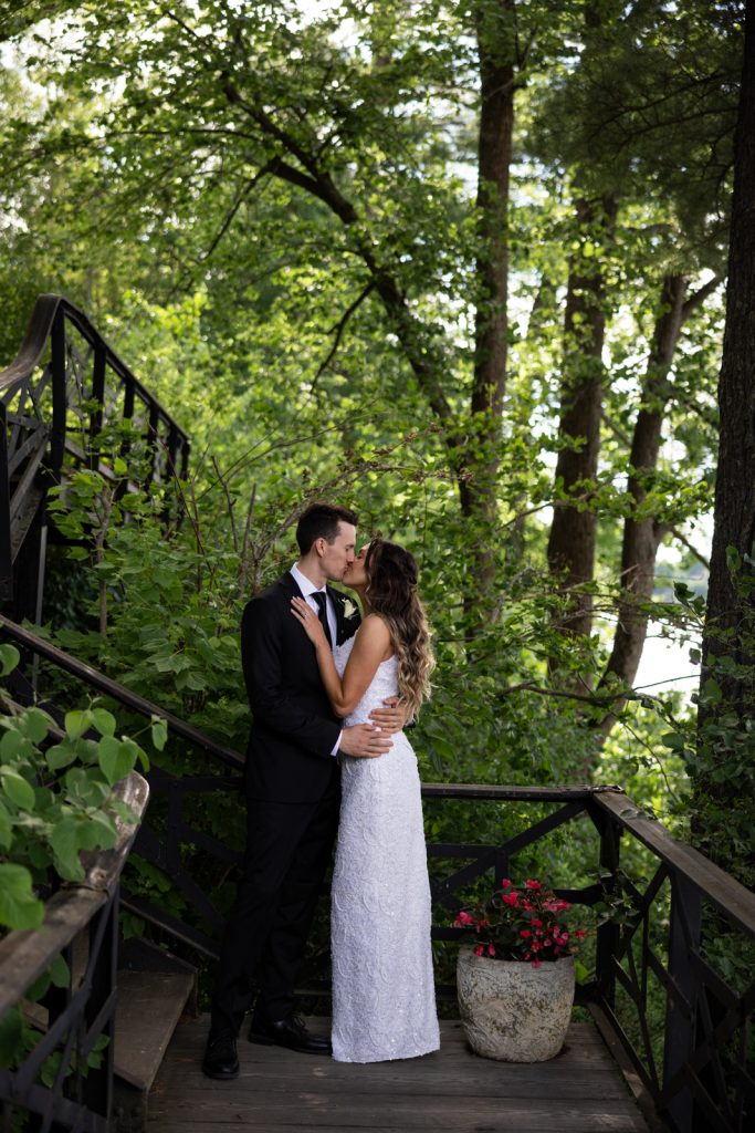 Brittany and Cole kissing on Stout’s Island wedding day, lakeside setting and lush greenery.