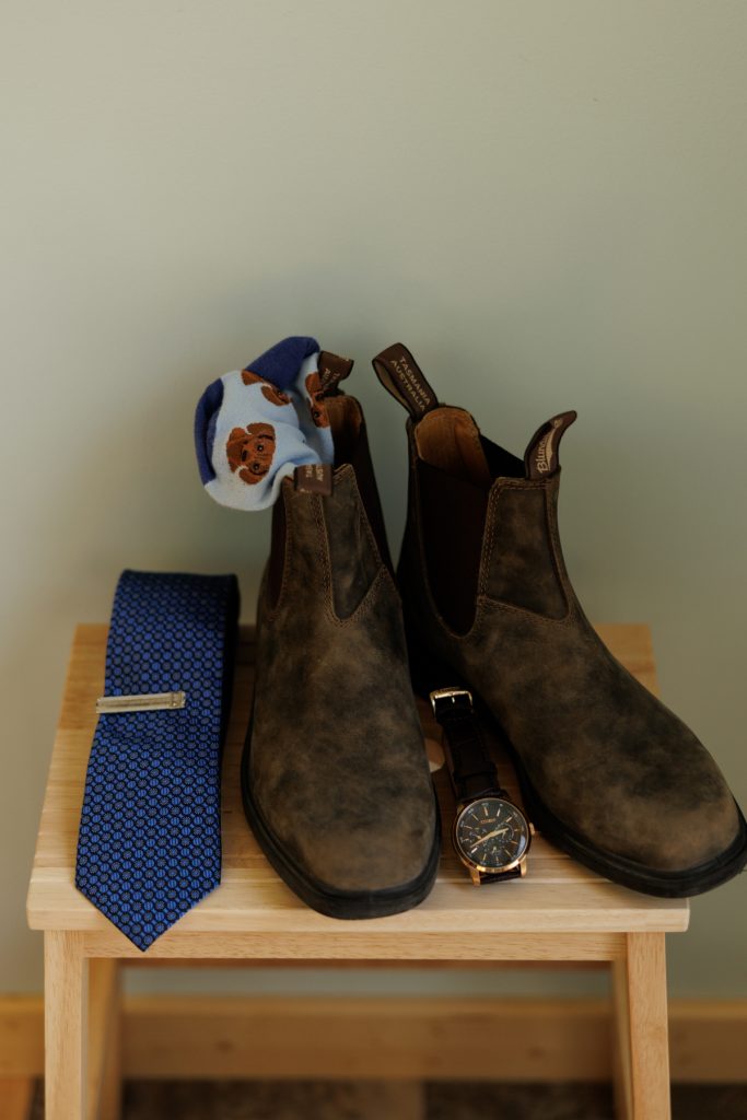 Groom's brown leather boots, socks, tie, and watch on wooden surface at Hayward, WI wedding.