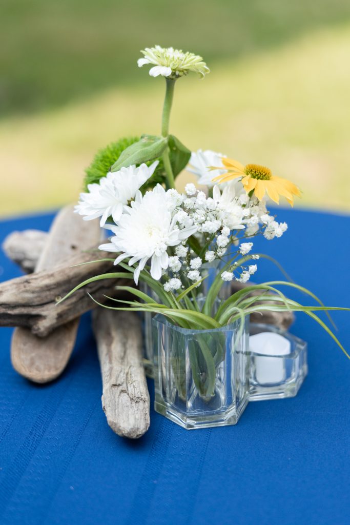 Rustic wedding centerpiece with white daisies and greenery on blue tablecloth and driftwood.