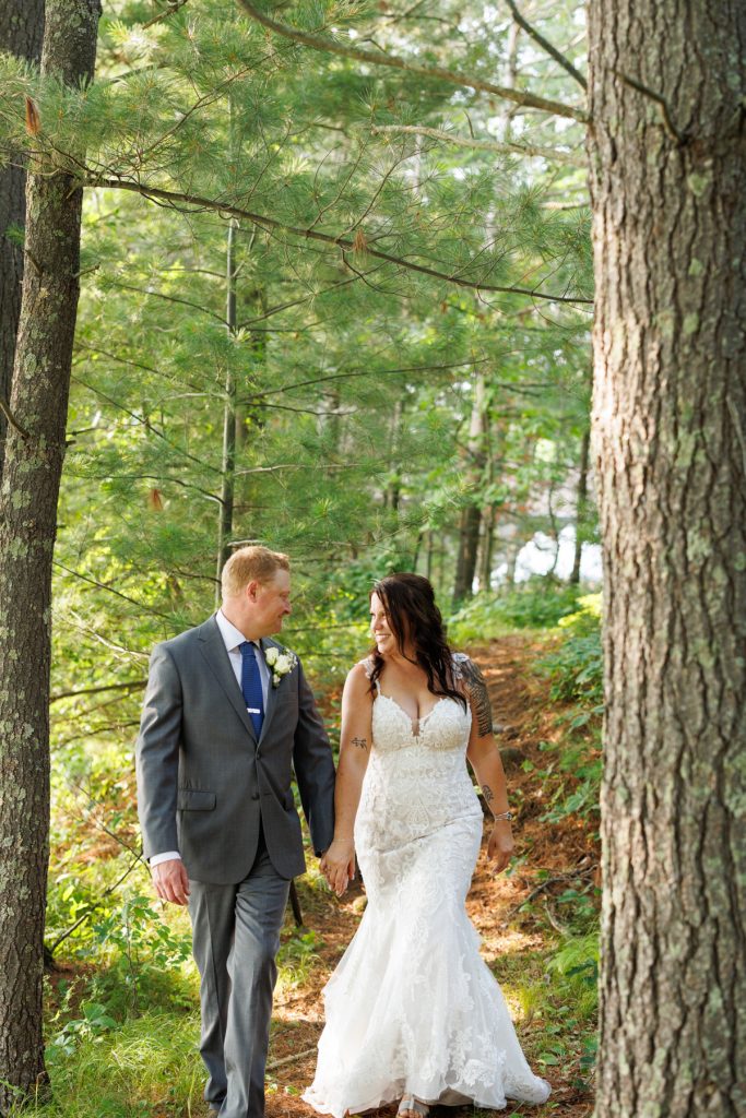 Bride and groom holding hands walking through lakeside trees at Hayward, WI wedding.