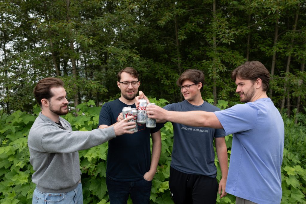 Four young men toasting with cans of beer outdoors, likely celebrating a wedding or special