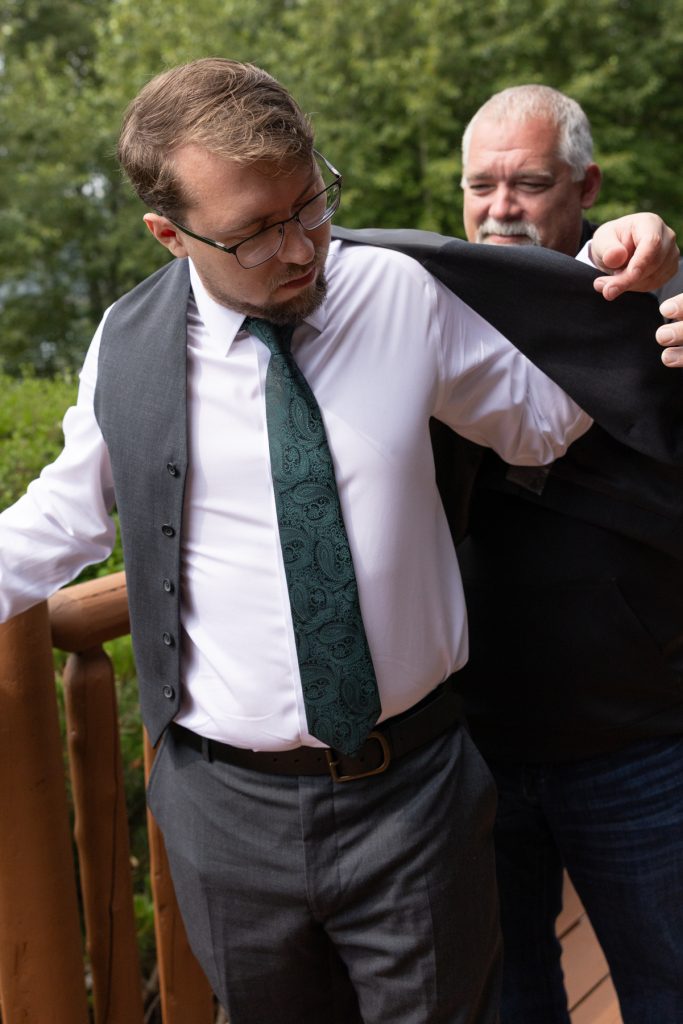 Groom in a gray suit and green tie adjusting his jacket with assistance