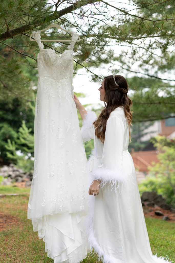 Bride in white robe admiring her wedding dress hanging from a tree branch, showcasing wedding