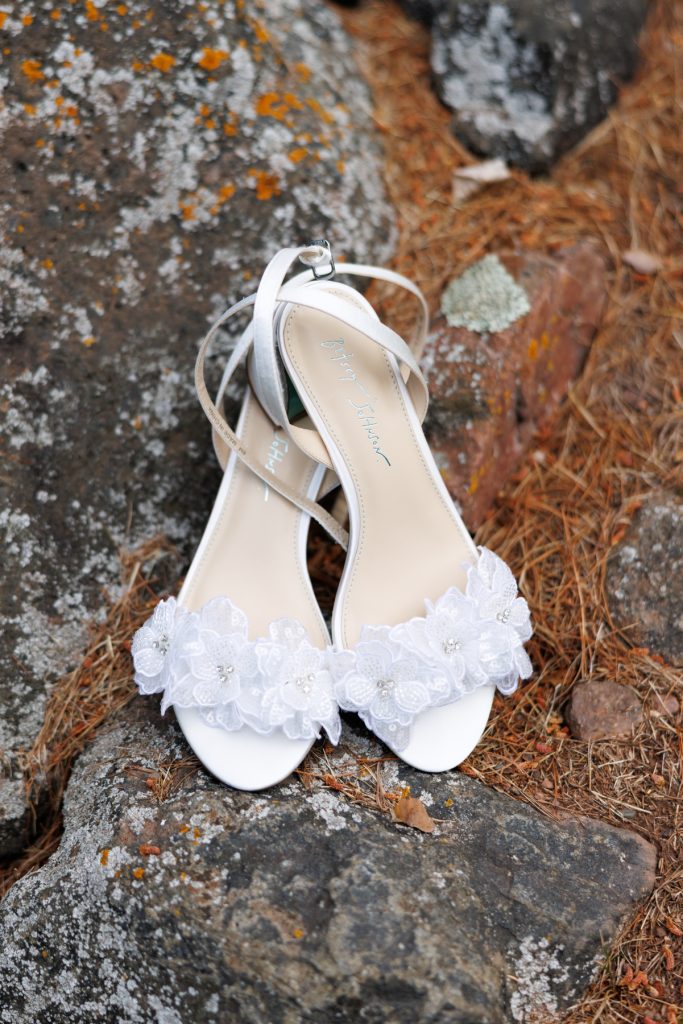 Elegant white wedding shoes with delicate floral details sit on a mossy rock, showcasing wedding