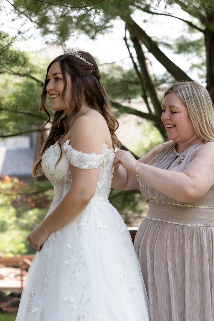 Bride in a detailed off-the-shoulder wedding dress smiles as her bridesmaid adjusts her gown amidst