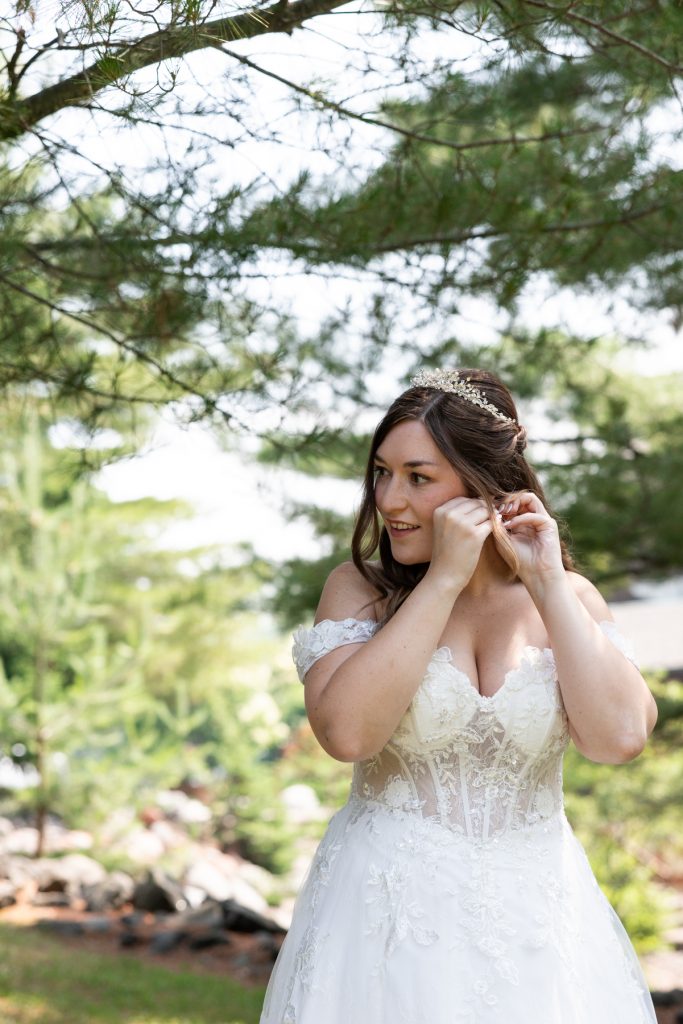 Bride in a detailed wedding dress adjusts her earrings in a wooded setting, showcasing wedding