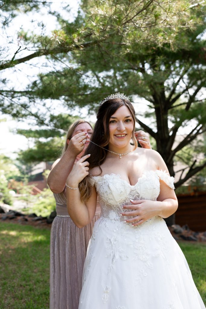 Bride in a white lace wedding dress smiles as a bridesmaid helps adjust her necklace