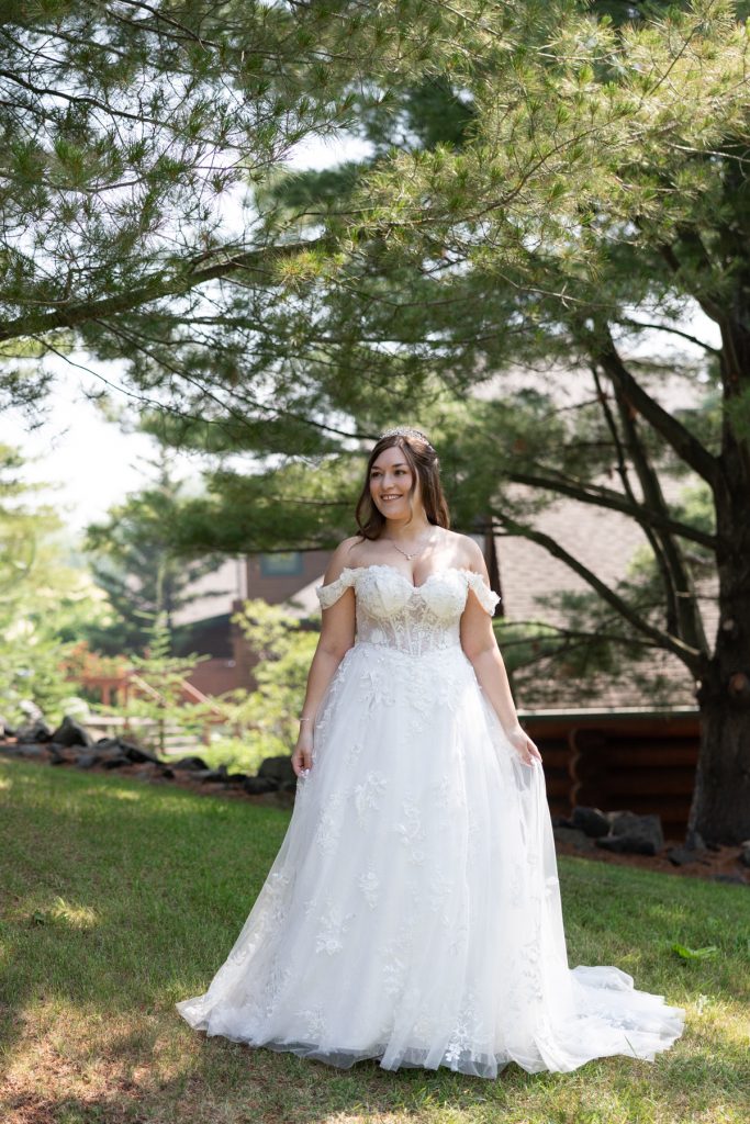 Bride in a flowing white wedding gown stands outdoors in a wooded setting, showcasing elegant