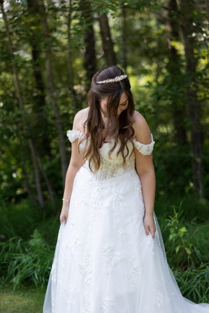 Bride in a white lace wedding dress and jeweled headband stands in a wooded area