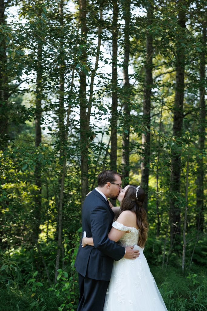 Bride and groom share a tender kiss embraced in a lush, wooded setting, showcasing romantic