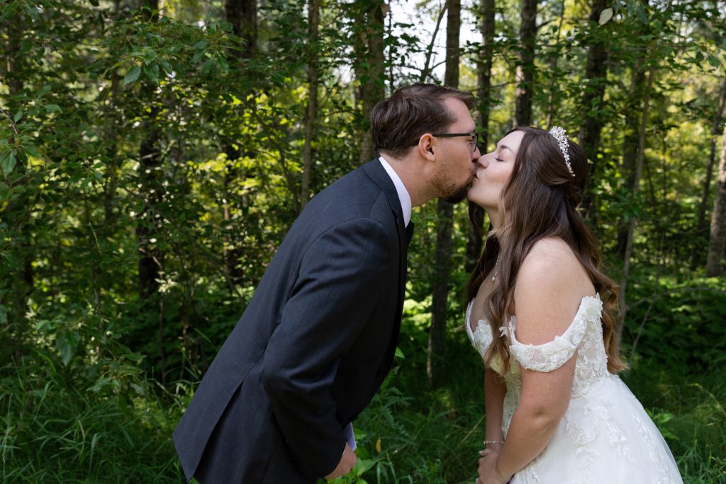 Bride and groom share a romantic kiss in a wooded setting, showcasing wedding photography services