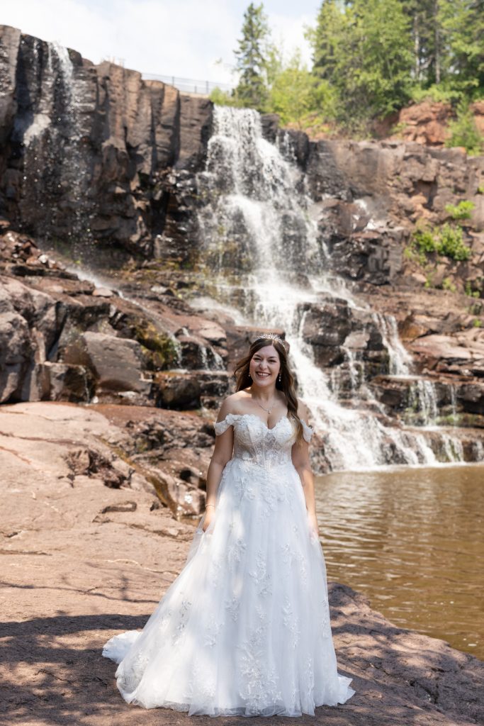 Bride in a flowing wedding dress smiles in front of a scenic waterfall in Wisconsin