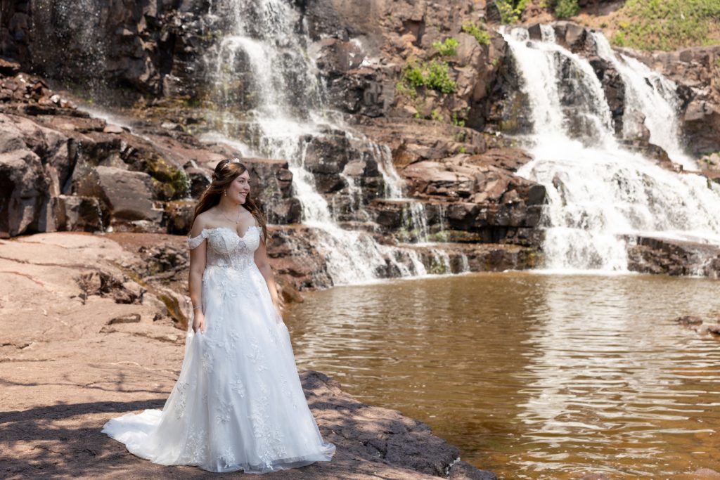Bride in a flowing white wedding dress stands near a cascading waterfall in Wisconsin, captured