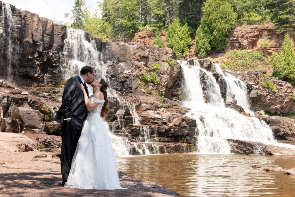 Bride and groom embracing in a romantic kiss near a cascading waterfall, showcasing wedding photography