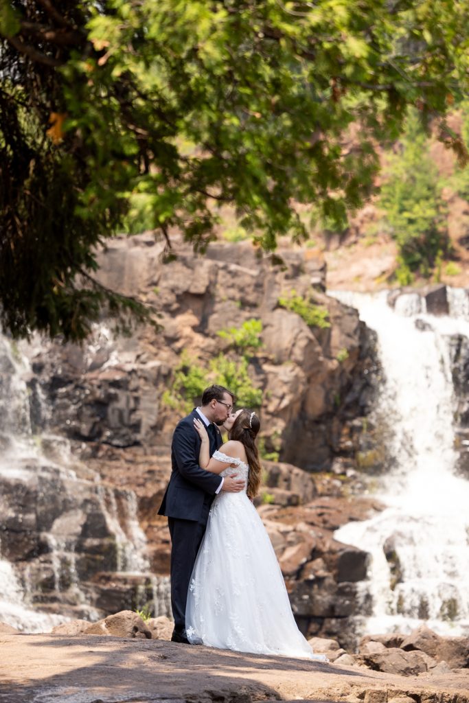 Bride and groom share a romantic kiss during their wedding day near a scenic waterfall