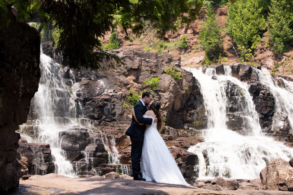 Bride and groom sharing a romantic kiss during their wedding day near a scenic waterfall