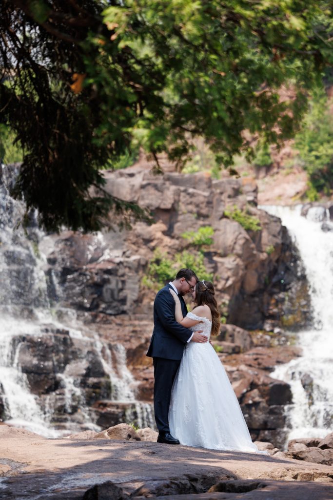 Bride and groom embracing in wedding attire near a scenic waterfall, showcasing romantic wedding photography