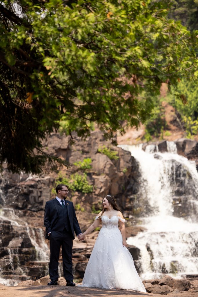 Bride and groom holding hands in wedding attire, posing for a photograph with a scenic