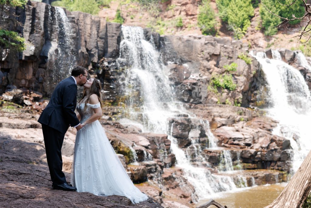 Bride and groom sharing a tender kiss near a cascading waterfall, captured by Beau Petersen