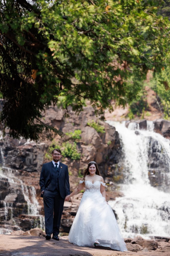 Newlyweds holding hands and walking together in Wisconsin, with a scenic waterfall and lush greenery
