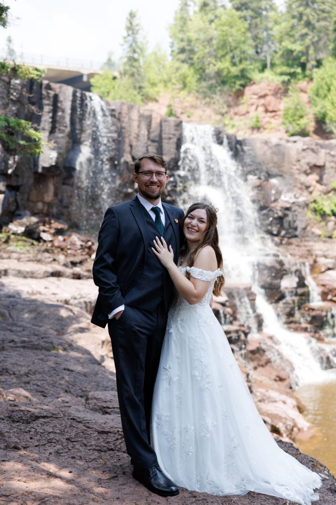 Newly married couple in formal wedding attire posing with a scenic waterfall backdrop, showcasing