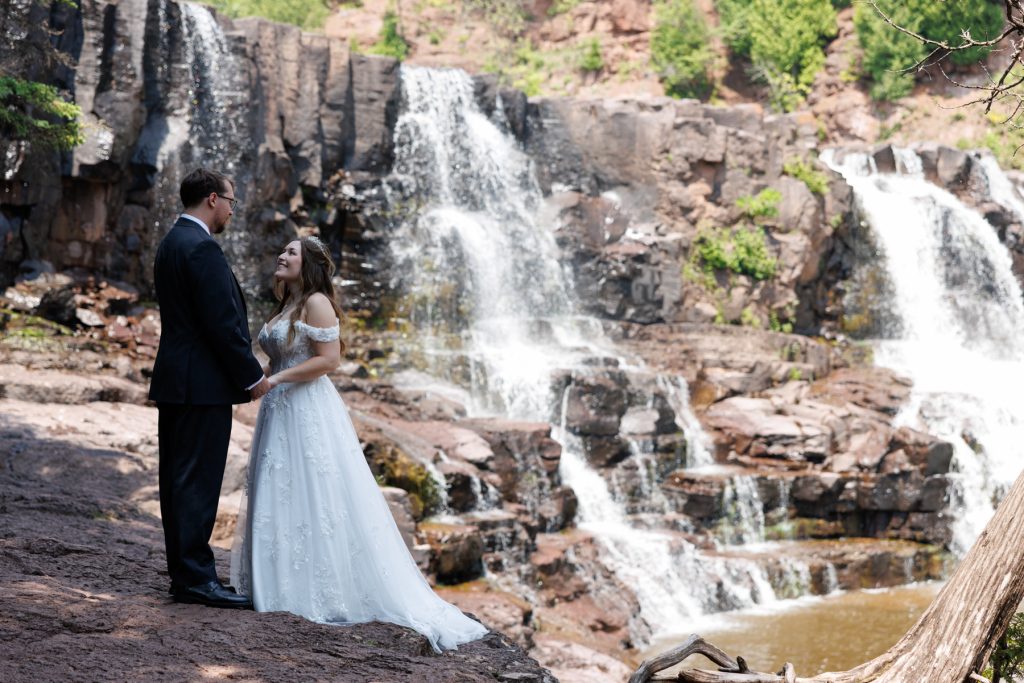 Newlyweds in formal wedding attire stand embracing near a scenic waterfall in Wisconsin, captured