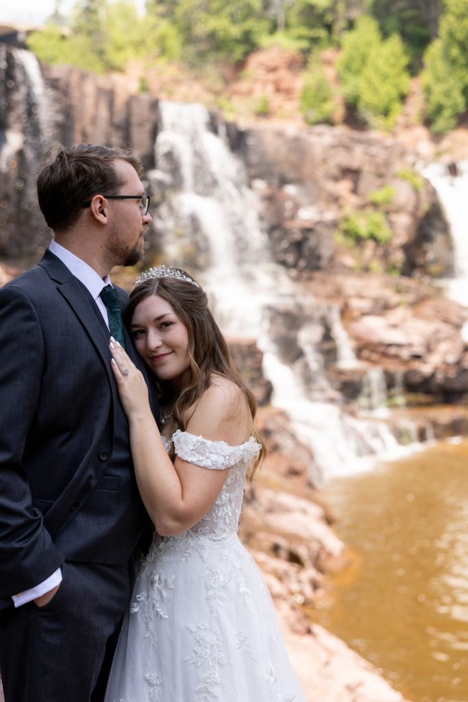 Bride and groom in wedding attire embracing near a scenic waterfall, captured by Beau Petersen
