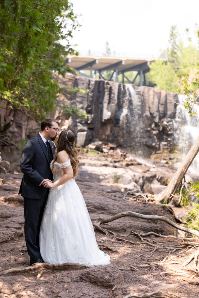 Bride and groom share a romantic kiss during their wedding day near a scenic waterfall