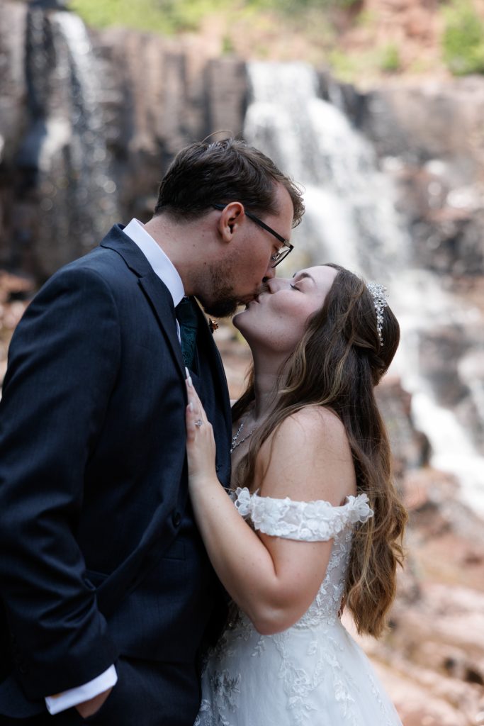 Bride and groom share a romantic kiss near a cascading waterfall, captured by Beau Petersen