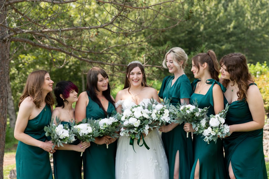 Bride surrounded by her bridesmaids in emerald green dresses, holding white floral bouquets, captured