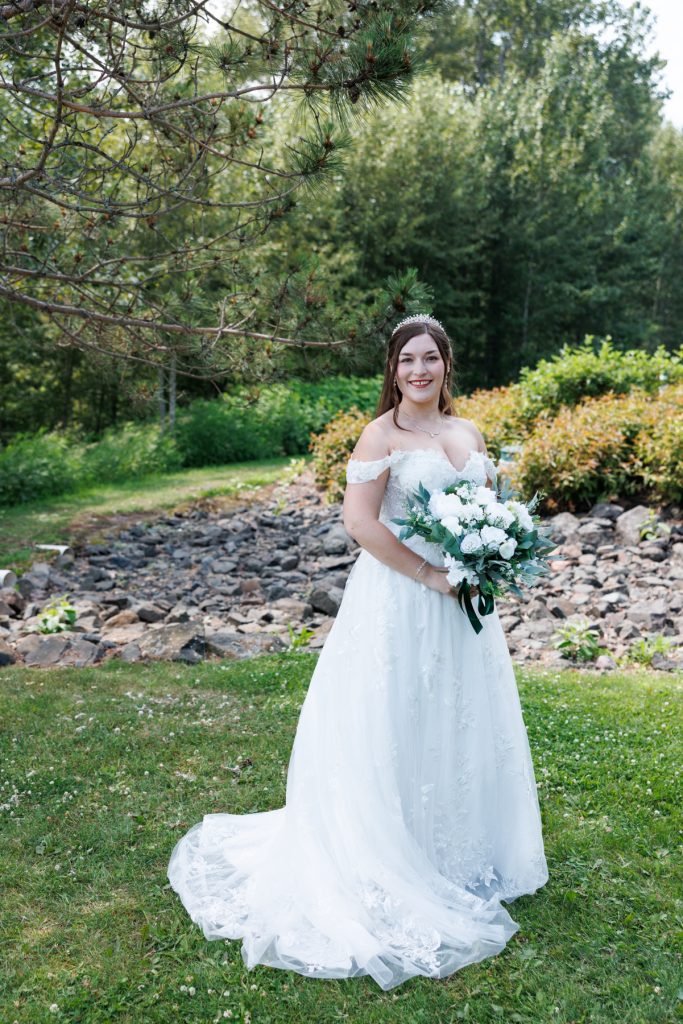 Bride in a white off-the-shoulder wedding dress and floral bouquet poses outdoors in a wooded