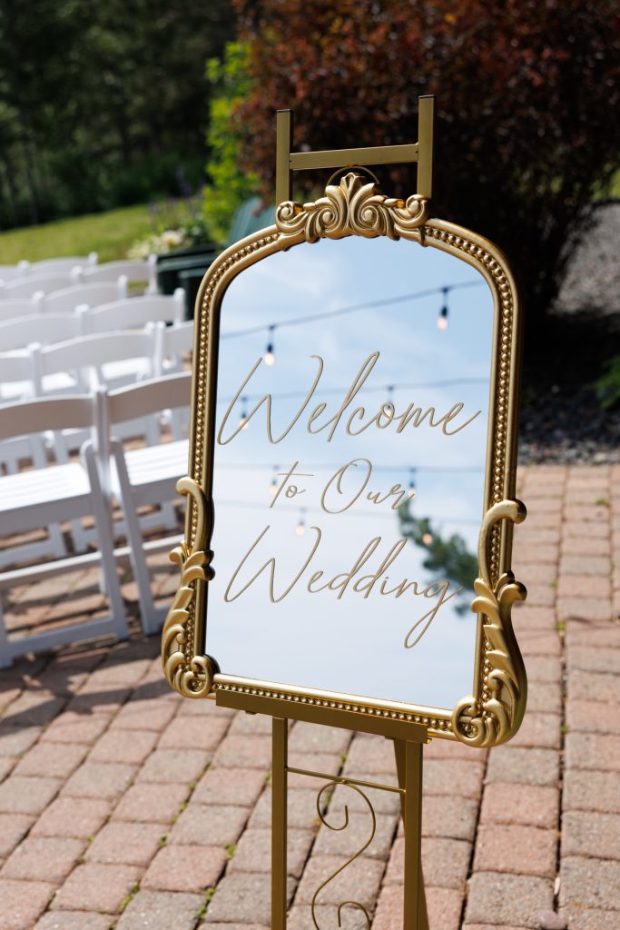 Elegant gold wedding welcome sign displayed outdoors, reflecting the sky and string lights, showcasing