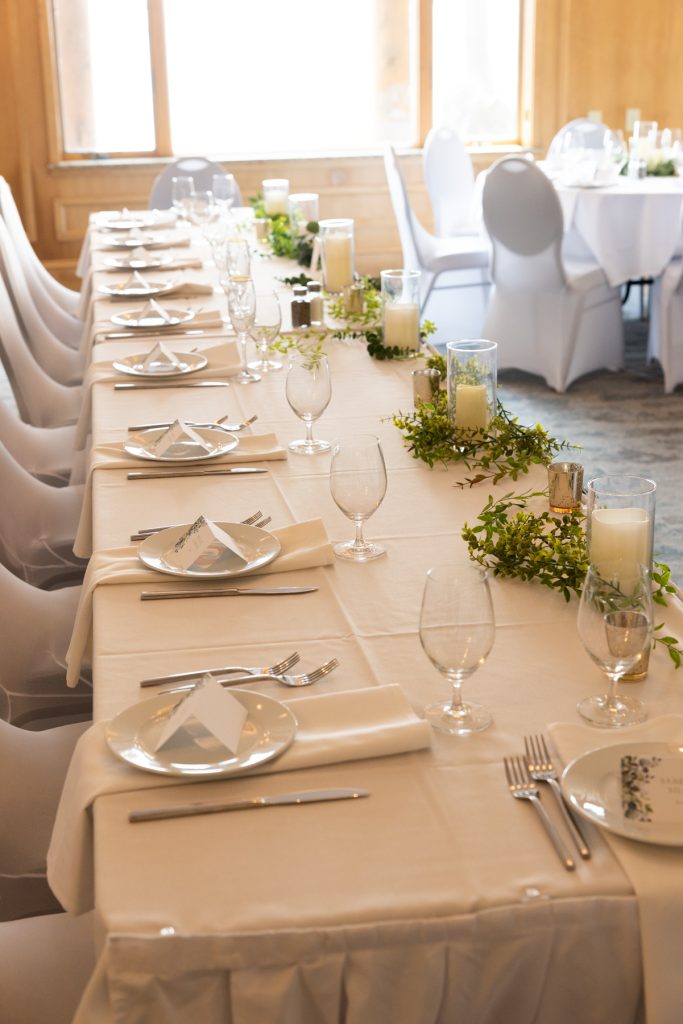 Long wedding reception table elegantly set with white linens, glassware, and greenery, showcasing Beau Petersen