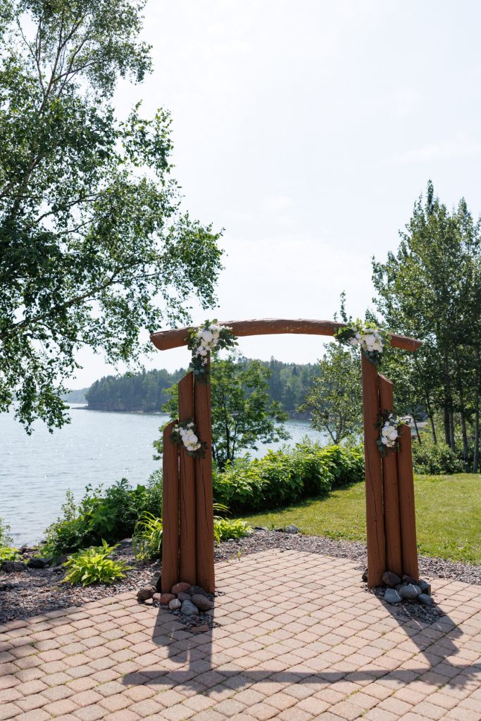Wooden wedding arch decorated with white flowers and greenery, overlooking a scenic lake and forest