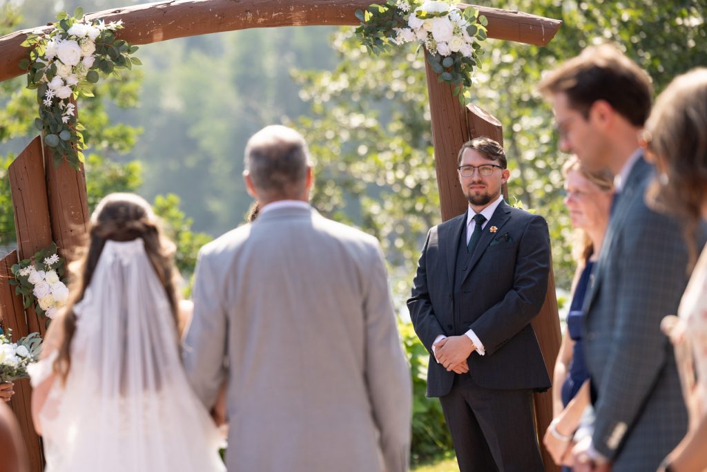 A groom in a dark suit stands attentively at a rustic wooden wedding arch decorated