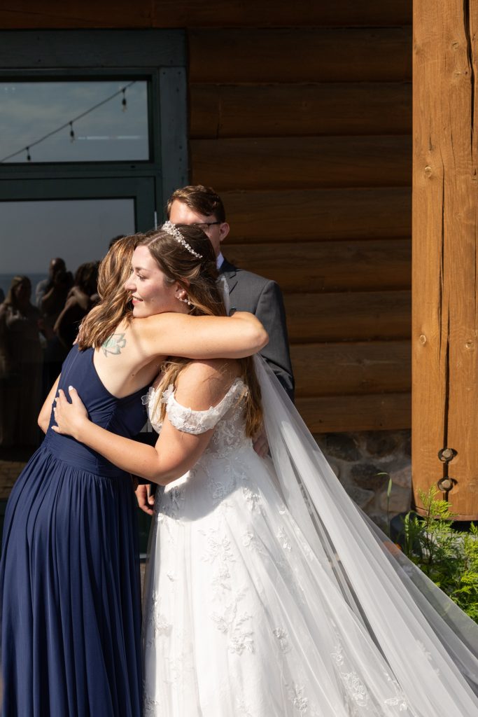 Bride in a white wedding dress hugs a bridesmaid in a blue dress, capturing