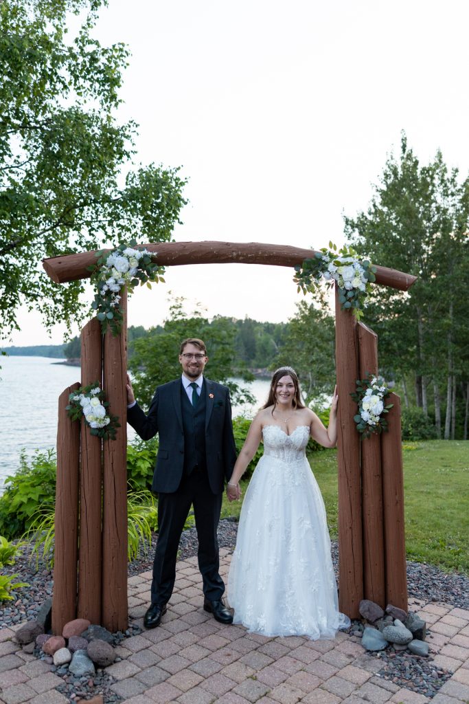 Newly married couple stands proudly holding hands beneath a rustic floral arch at their wedding