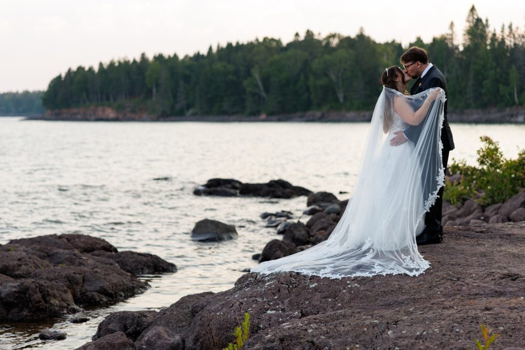 Bride and groom share a romantic kiss on the rocky shoreline of a lake, captured