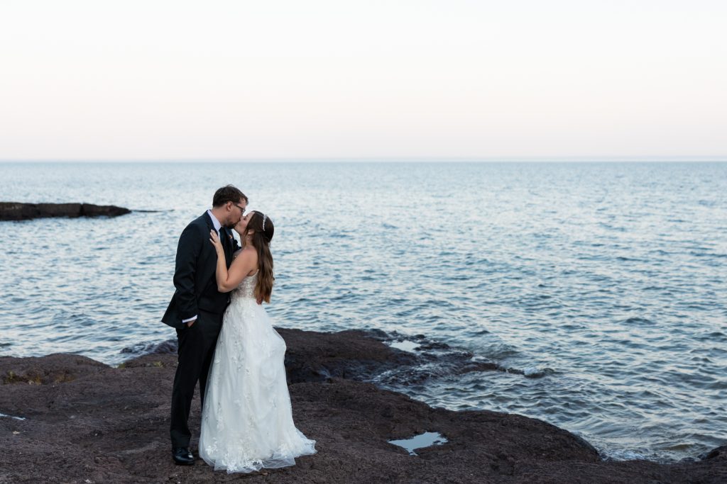 Bride and groom embracing in wedding attire against a backdrop of Lake Superior, showcasing romantic