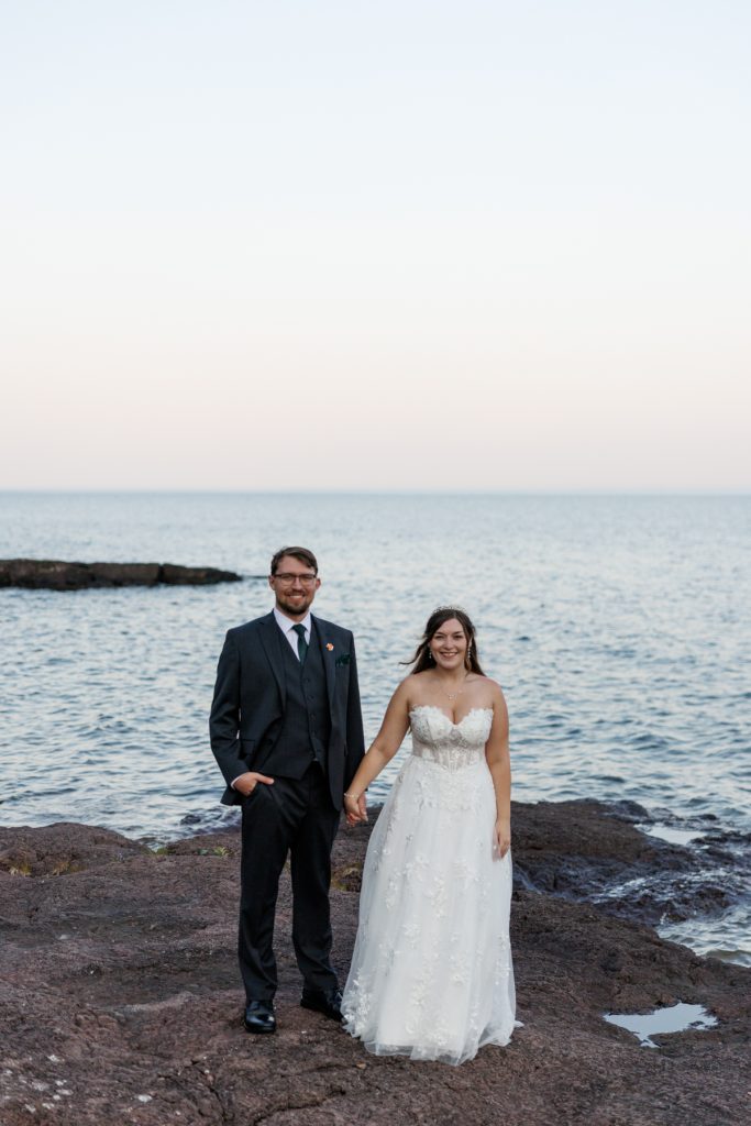 Newly married couple holding hands, smiling, with Lake Superior as a scenic backdrop, showcasing Wisconsin